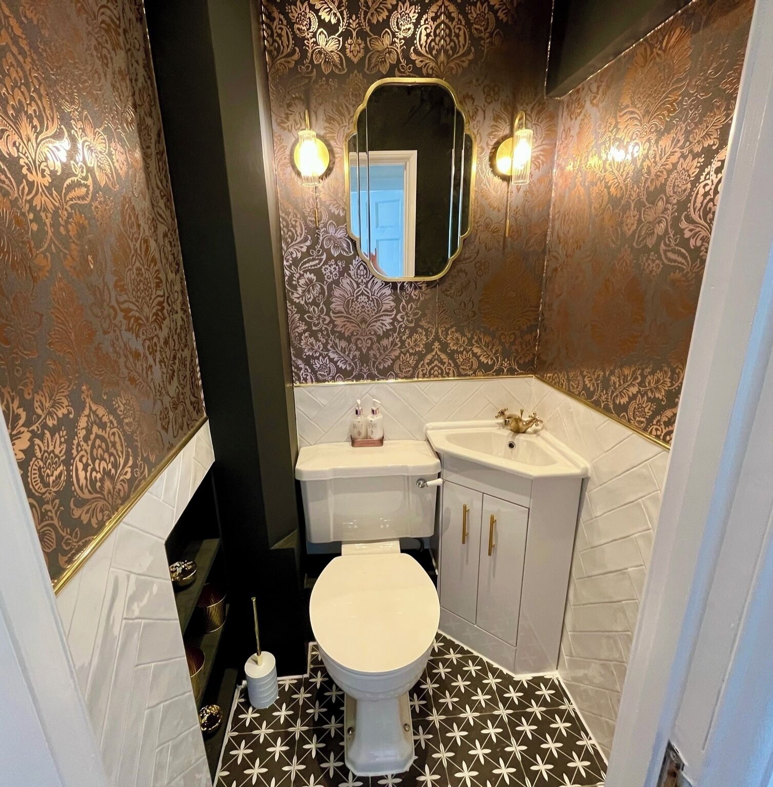 A bathroom with a white toilet, a sink with gold handles, and patterned wallpaper.