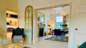 A spacious living room with a wooden coffee table, a green sofa, and a bookshelf. An open door leads to a dining area with a chandelier. - The Paragon Penthouse - Stunning Views over Bath! (Bath)