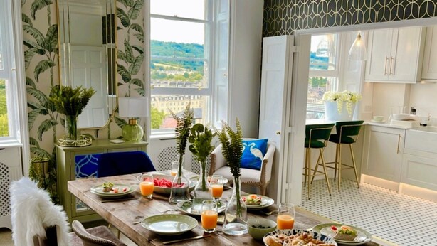 A dining area with a wooden table set for breakfast, a view of the city through large windows, and a kitchen with white cabinets and a patterned backsplash. - The Paragon Penthouse - Stunning Views over Bath! (Bath)