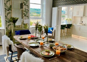 Spiseområde med et træbord dækket til morgenmad, udsigt til byen gennem store vinduer og et køkken med hvide skabe og et mønstret bagpanel.