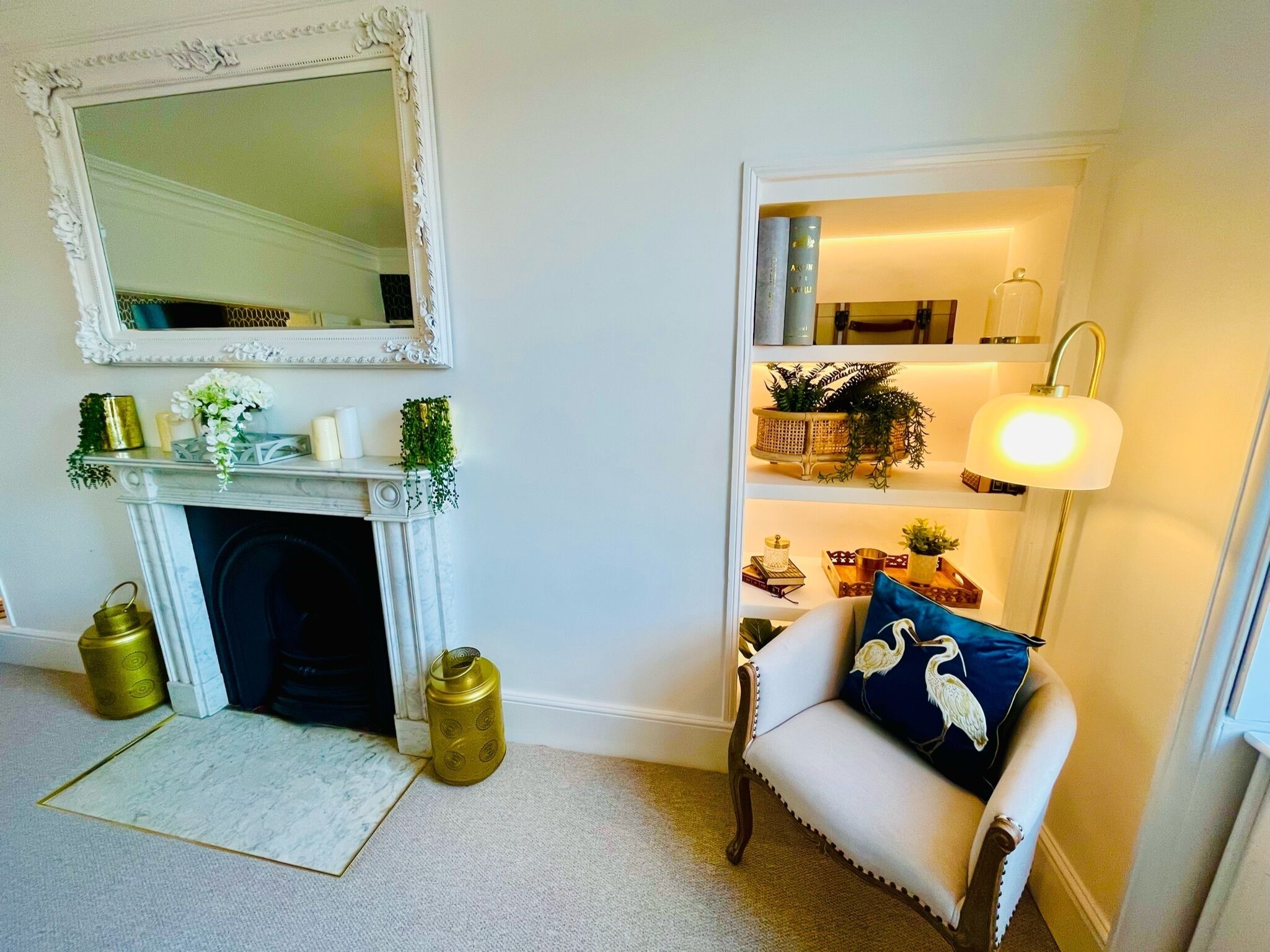 A room with a white fireplace, a mirror, a shelf with decorative items, and a chair with a blue pillow.