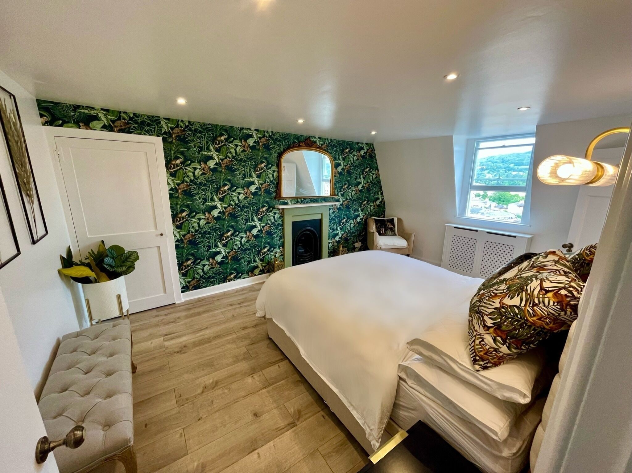 A bedroom with a bed, a chair, a small table with plants, a fireplace, and a window with a view.