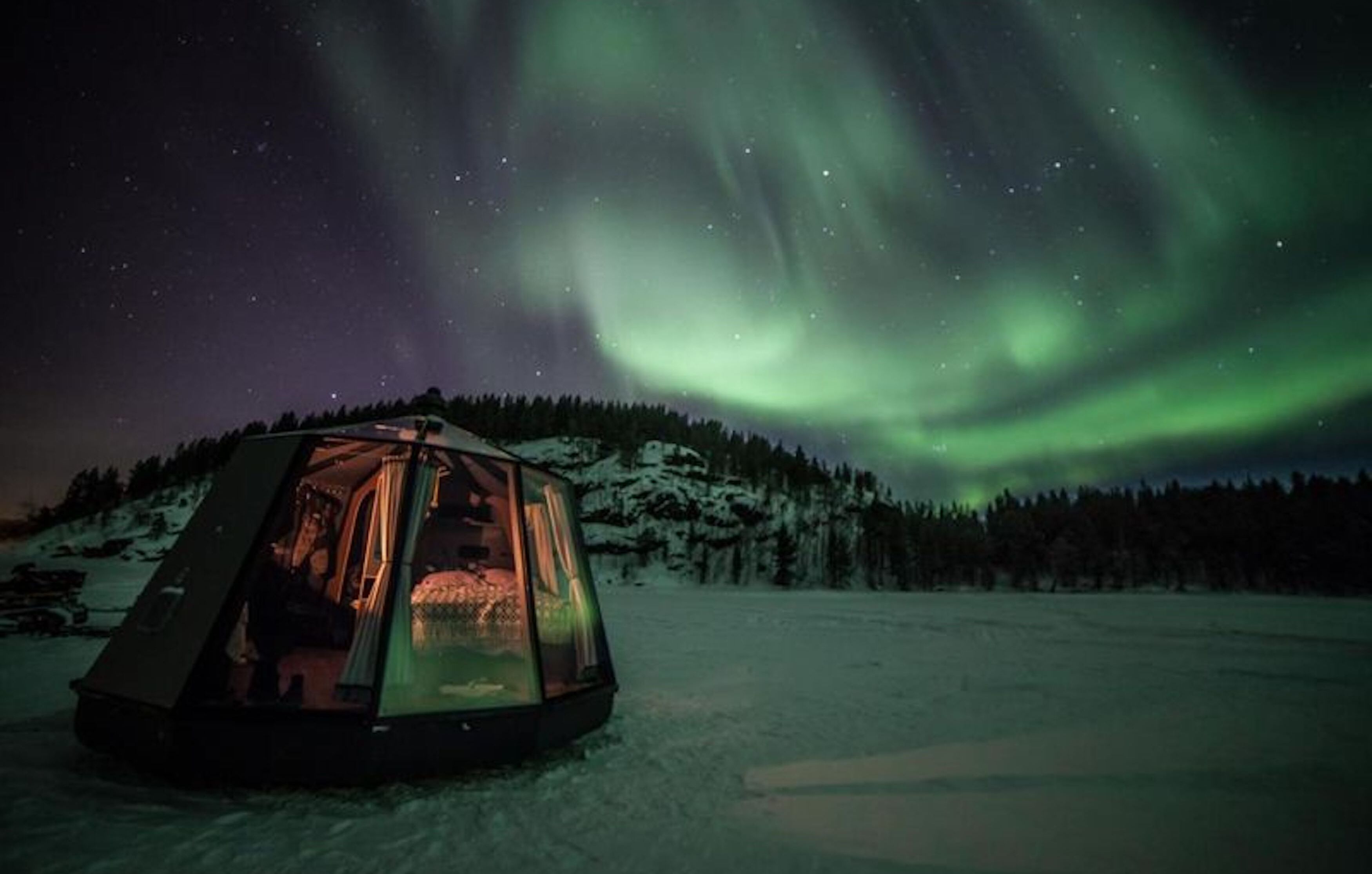 Een geodetische koepeltent met daarin een persoon, gelegen in een besneeuwd landschap met een beboste heuvel op de achtergrond en het Noorderlicht erboven.