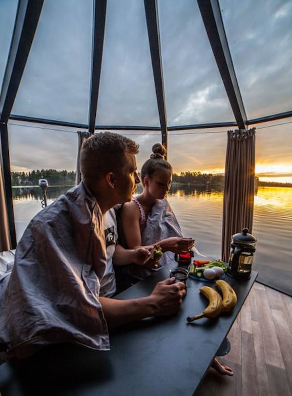 Pariskunta illastaa pöydässä, josta avautuu näkymä järvelle ja auringonlaskuun.