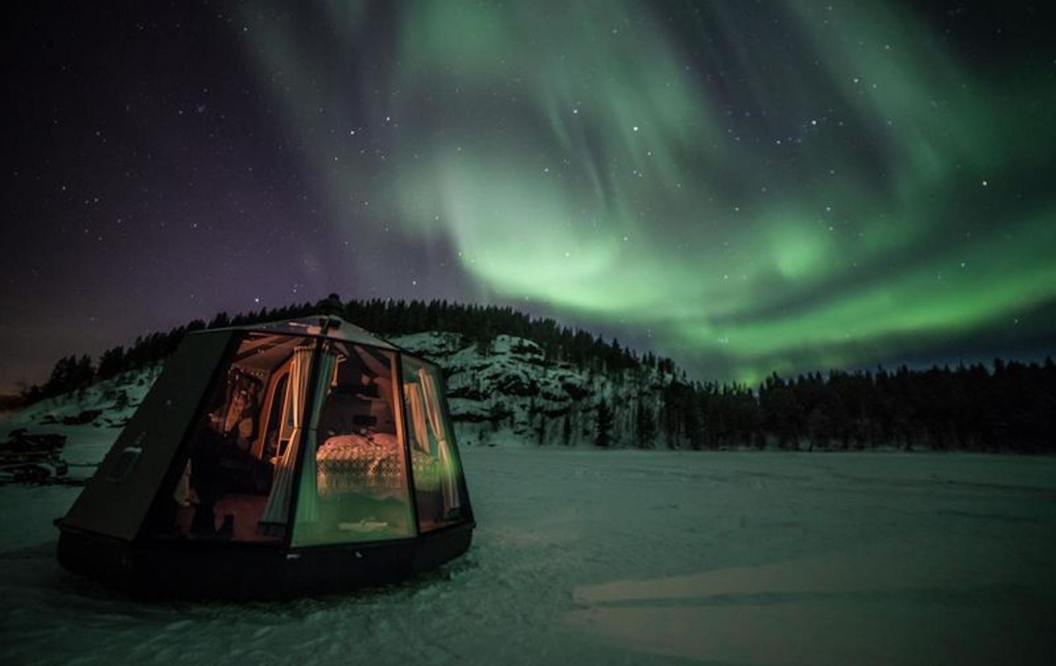 Ein geodätisches Kuppelzelt mit einer Person darin, auf einer verschneiten Landschaft. Im Hintergrund ist ein bewaldeter Hügel zu sehen und über dem Zelt erstrecken sich die Polarlichter.