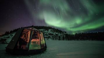 Ein geodätisches Kuppelzelt mit einer Person darin, auf einer verschneiten Landschaft. Im Hintergrund ist ein bewaldeter Hügel zu sehen und über dem Zelt erstrecken sich die Polarlichter.