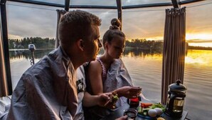 Una pareja cenando dentro de una cabaña con vista a un lago y al atardecer.