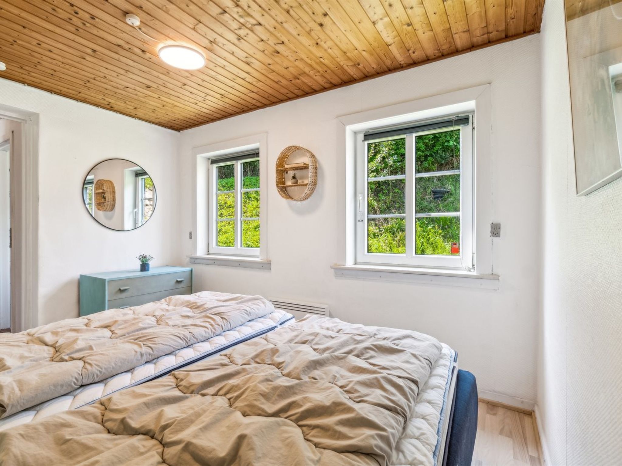 A bedroom with a wooden ceiling, two windows with greenery outside, a bed with a beige duvet, a blue dresser, and a round mirror on the wall.