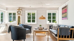 Ein geräumiges Wohnzimmer mit Holzbalkendecke, großen Fenstern, einer Uhr und einem modernen Sofa.