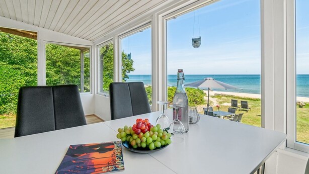 Ein Essbereich mit einem Tisch für zwei Personen, einer Wasserflasche und einer Schüssel mit Trauben. Große Fenster bieten Blick auf Strand und Meer.