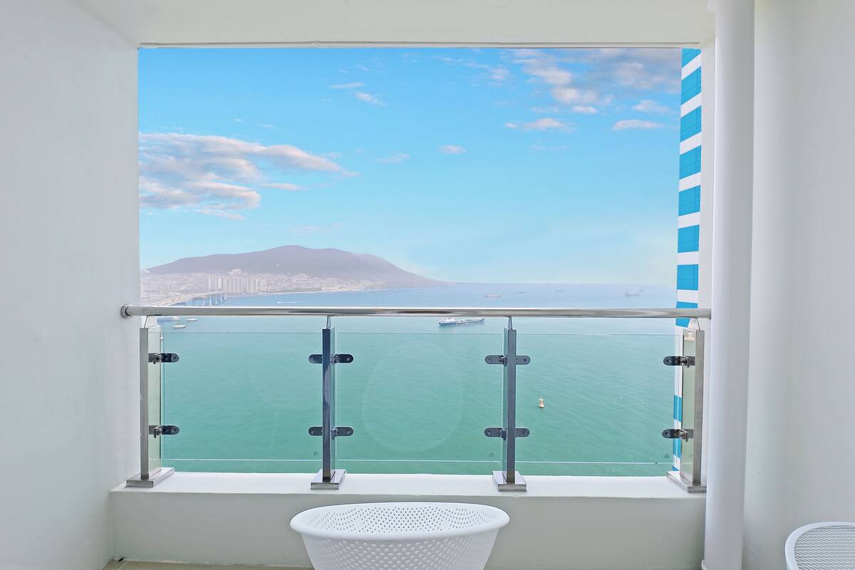 A balcony with a clear view of the sea, a distant mountain, and a cityscape.