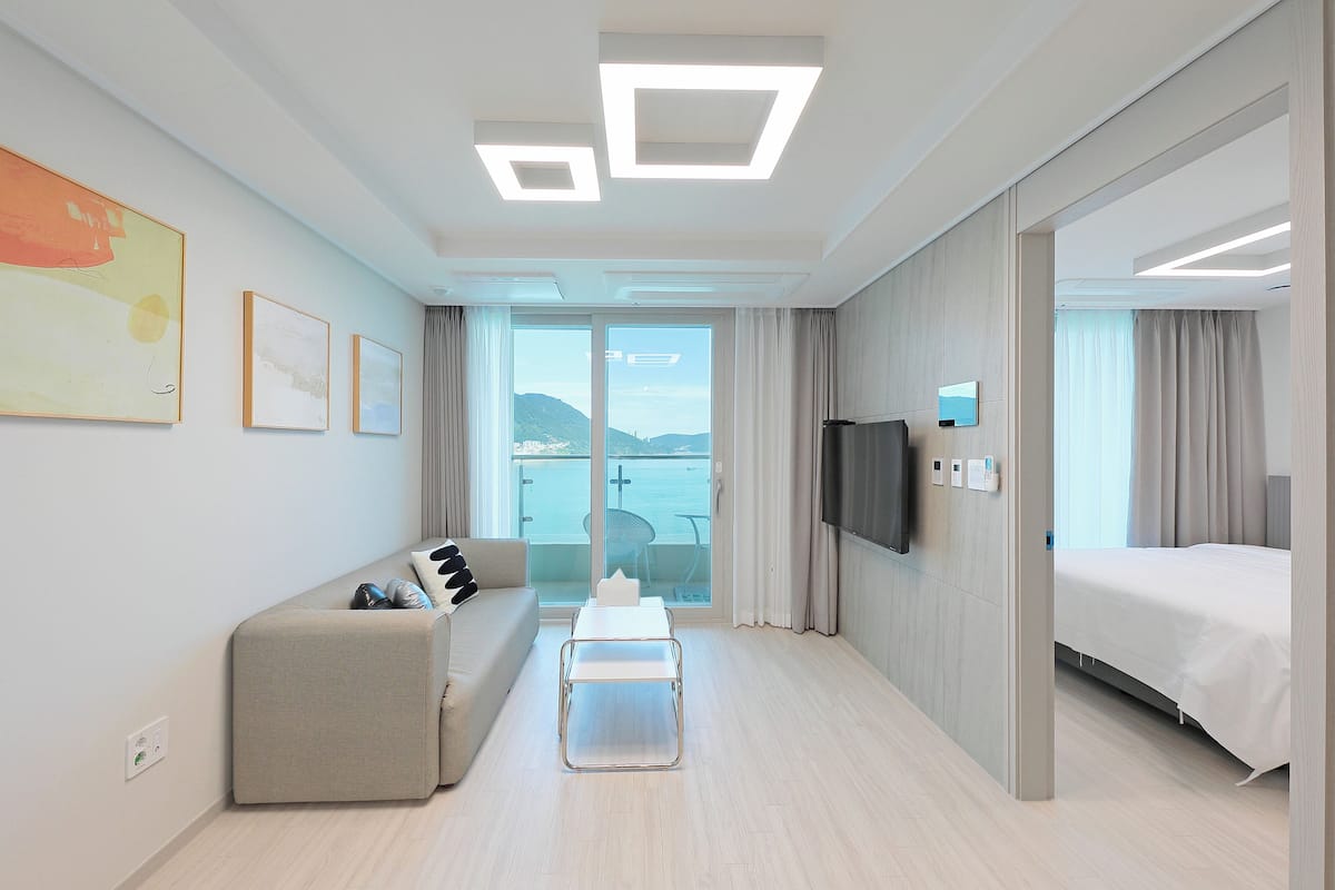 A modern hotel room with a sofa, a small table, a bed, a TV, and a view of the outdoors.