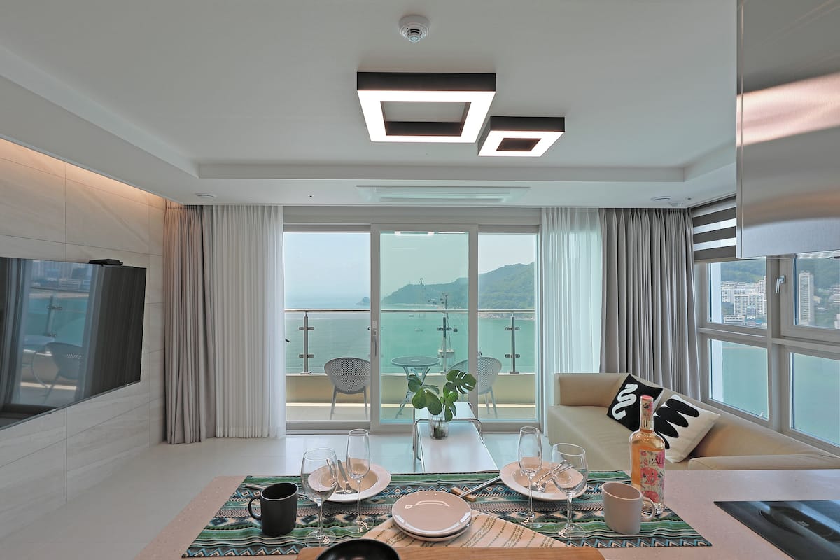 A modern hotel room with a balcony overlooking the sea, a dining area with table set for two, and a large flat-screen TV mounted on the wall.