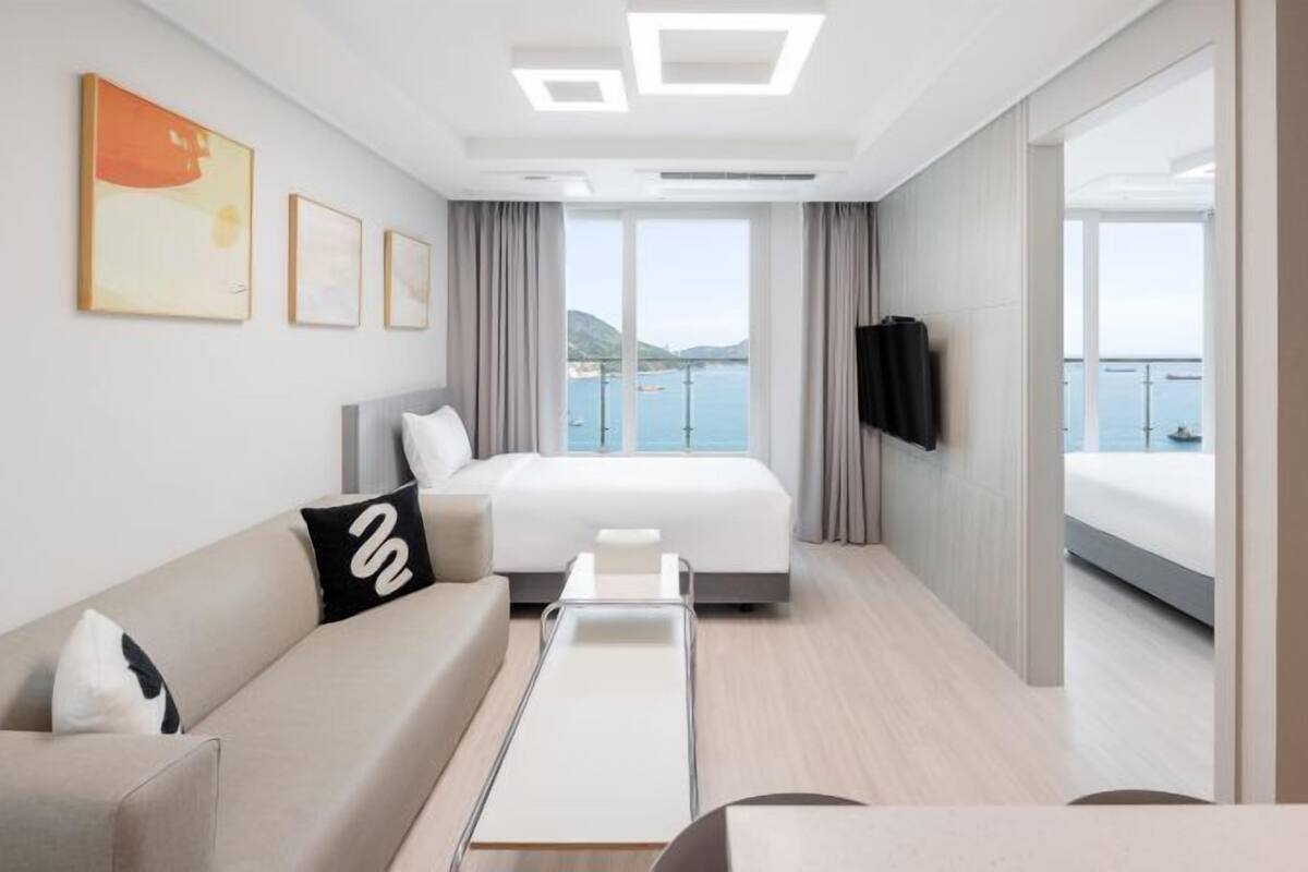 A modern hotel room with a bed, a sofa, a small table, and a TV. The room has large windows with a view of the sea.