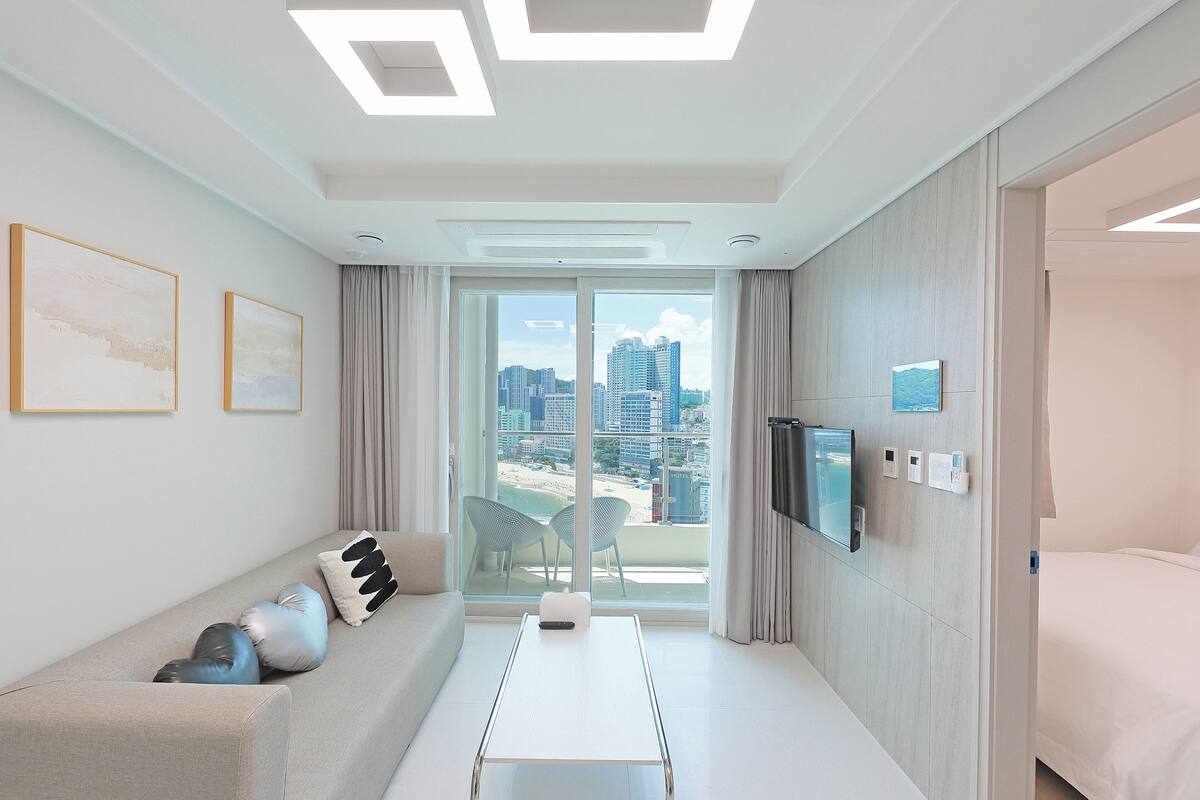 A modern hotel room with a sofa, a coffee table, a TV mounted on the wall, and a large window offering a city view.