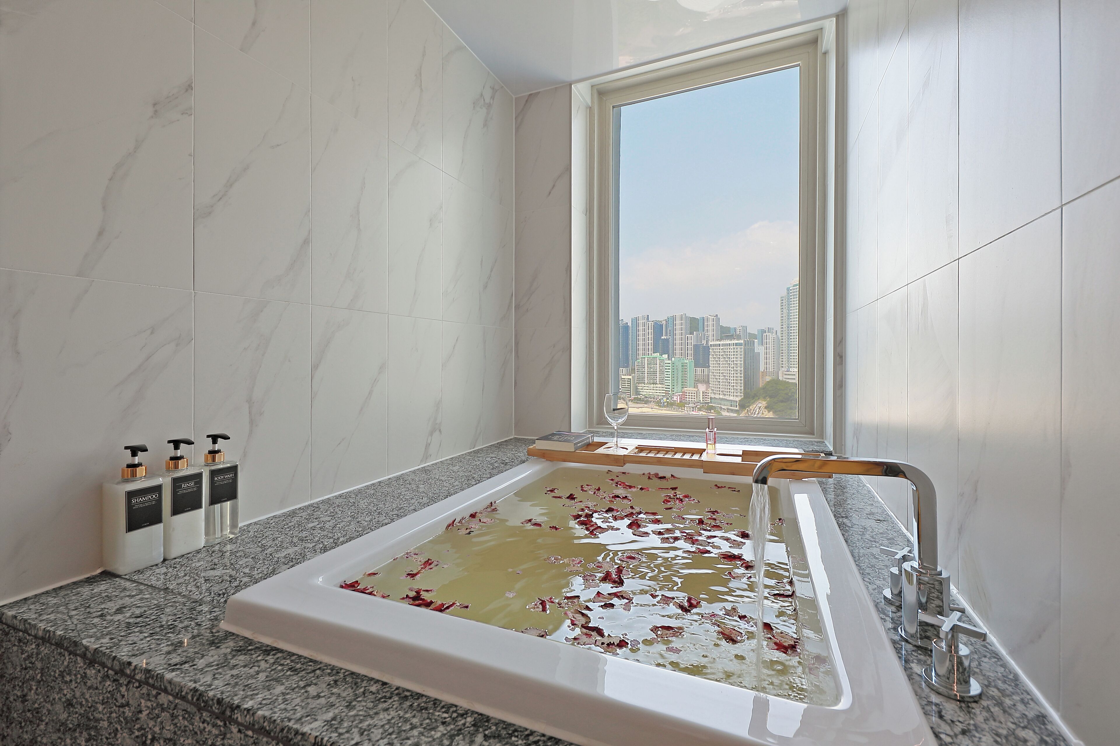 Een moderne badkamer met een groot bad vol water en rozenblaadjes, een raam met uitzicht op de stad, en toiletartikelen op het aanrecht.