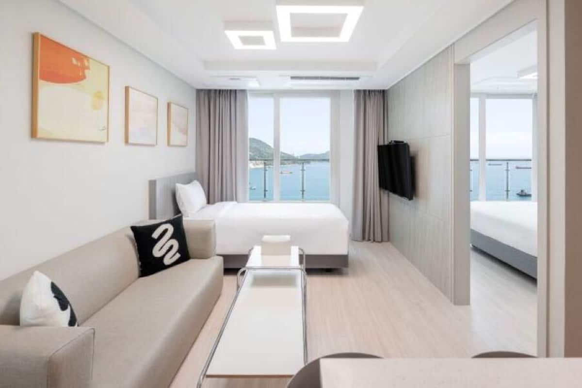 A modern hotel room with a bed, a sofa, a TV, and a balcony with a view of the sea.