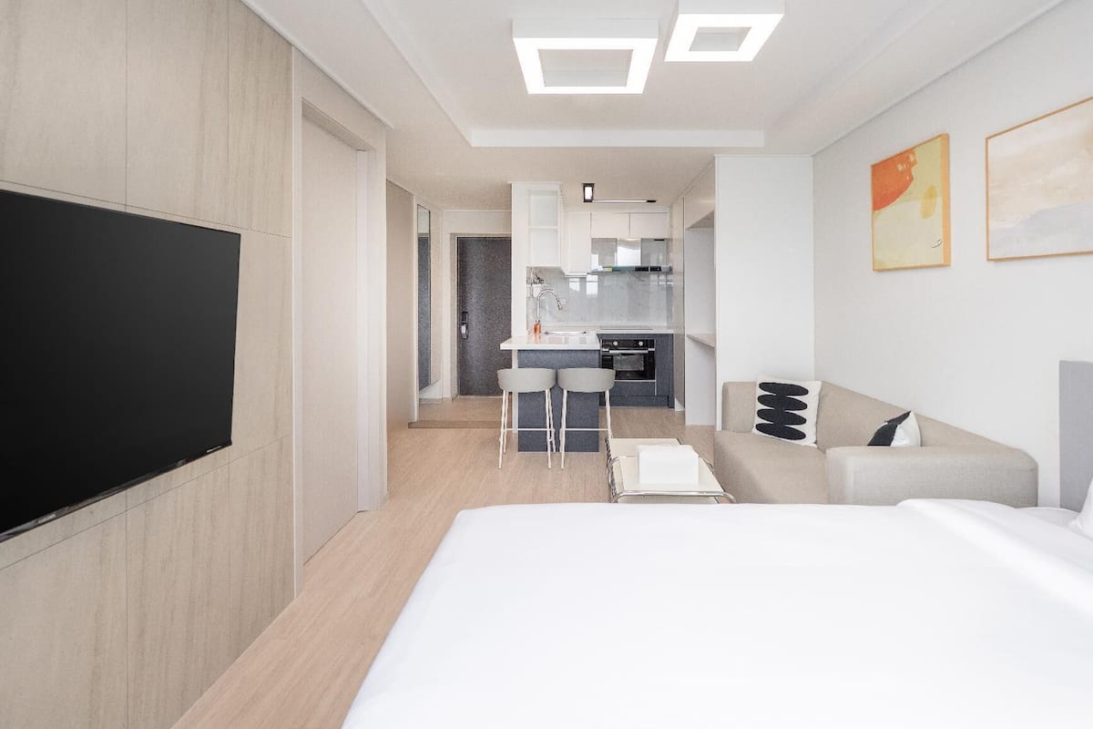 A modern hotel room with a flat-screen TV, a bed, a sofa, a small kitchen area, and a dining table.