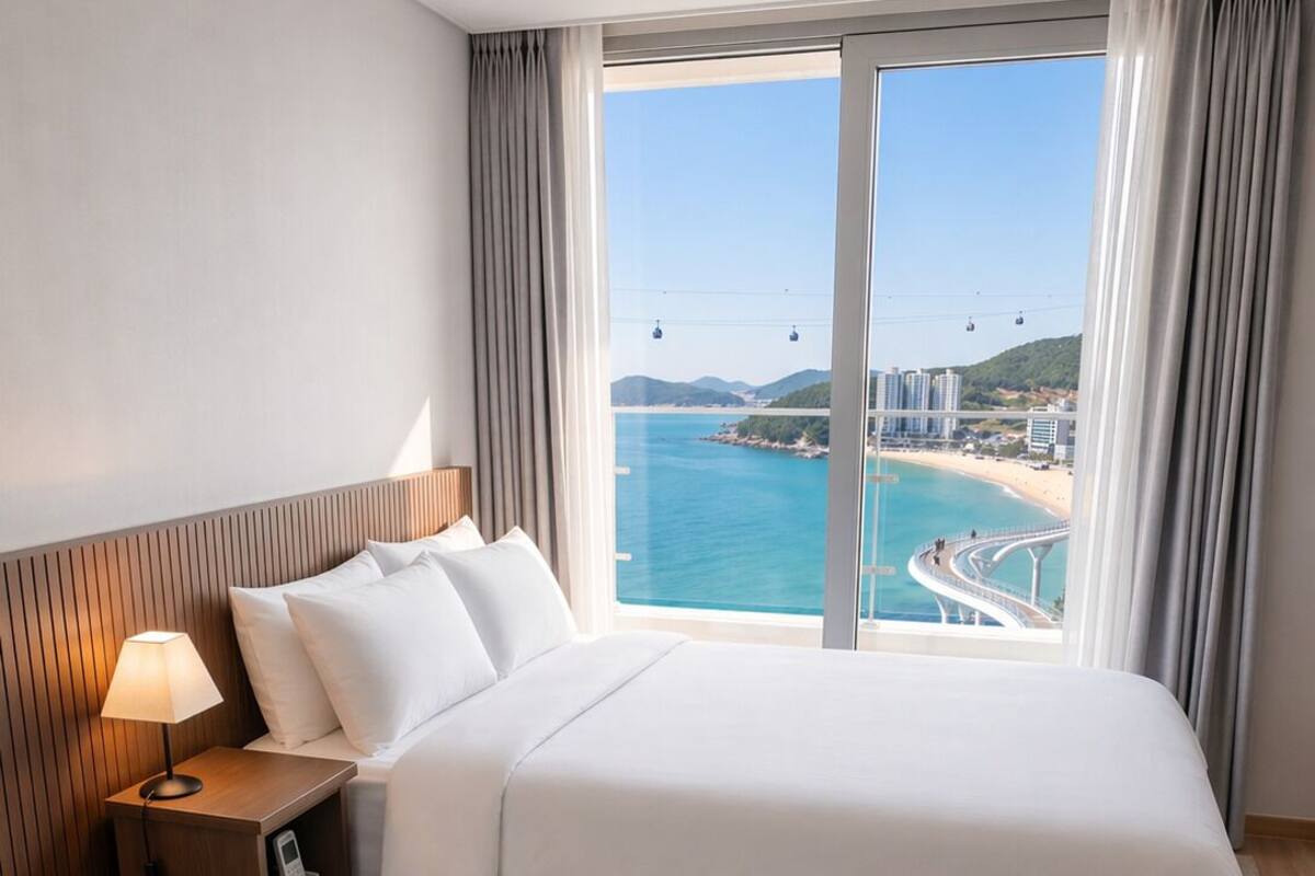 A hotel room with a bed, a bedside table with a lamp, a control panel, and a view of a beach and water.