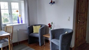 A room with a wooden ceiling, a window with curtains, two grey armchairs, a small table, and a wooden chair.