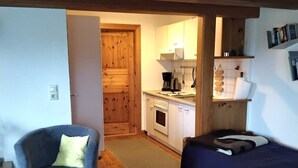 A room with a bed, a chair, a small table, a kitchenette, and a wooden door.