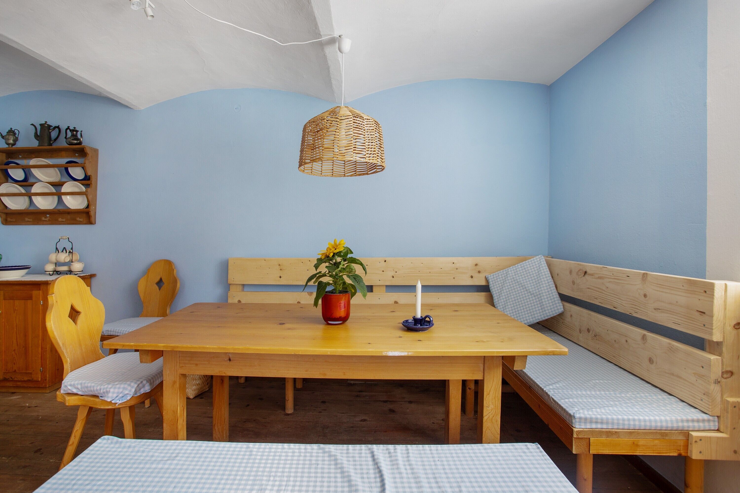 Une pièce avec une table en bois, des bancs, un luminaire suspendu et une étagère fixée au mur où sont rangés de la vaisselle.