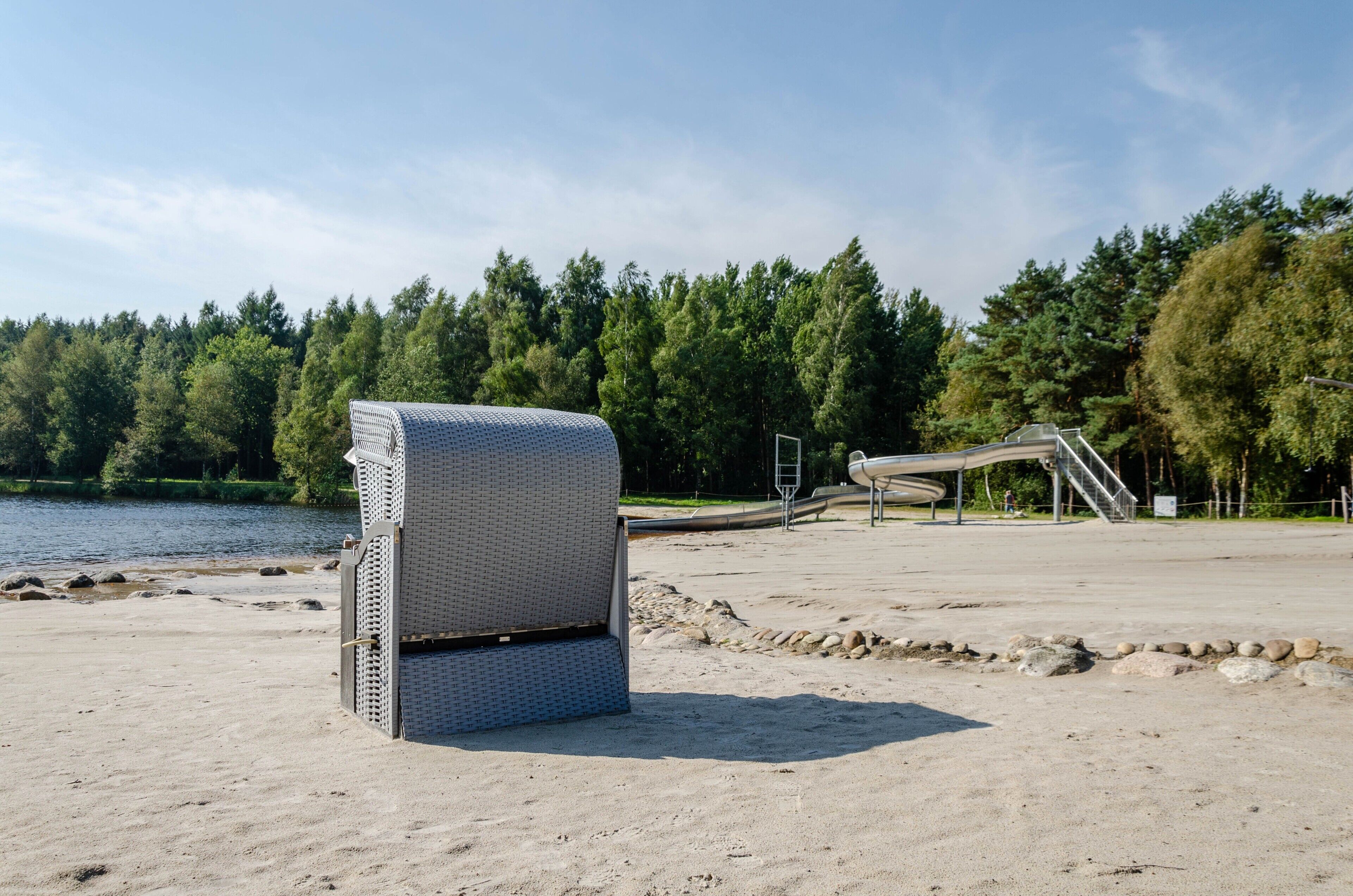 En strand med et overdækket siddeområde, en trappe op til en overdækket struktur og en skov i baggrunden.