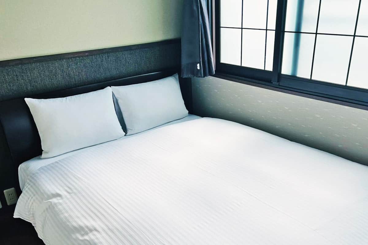 Cama arrumada com cabeceira escura e roupa de cama branca em quarto com janela e tomada na parede.