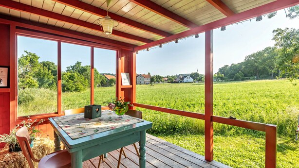 Un espace extérieur abrité, doté d’une table et de chaises, de grandes fenêtres et offrant une vue sur un champ verdoyant.
