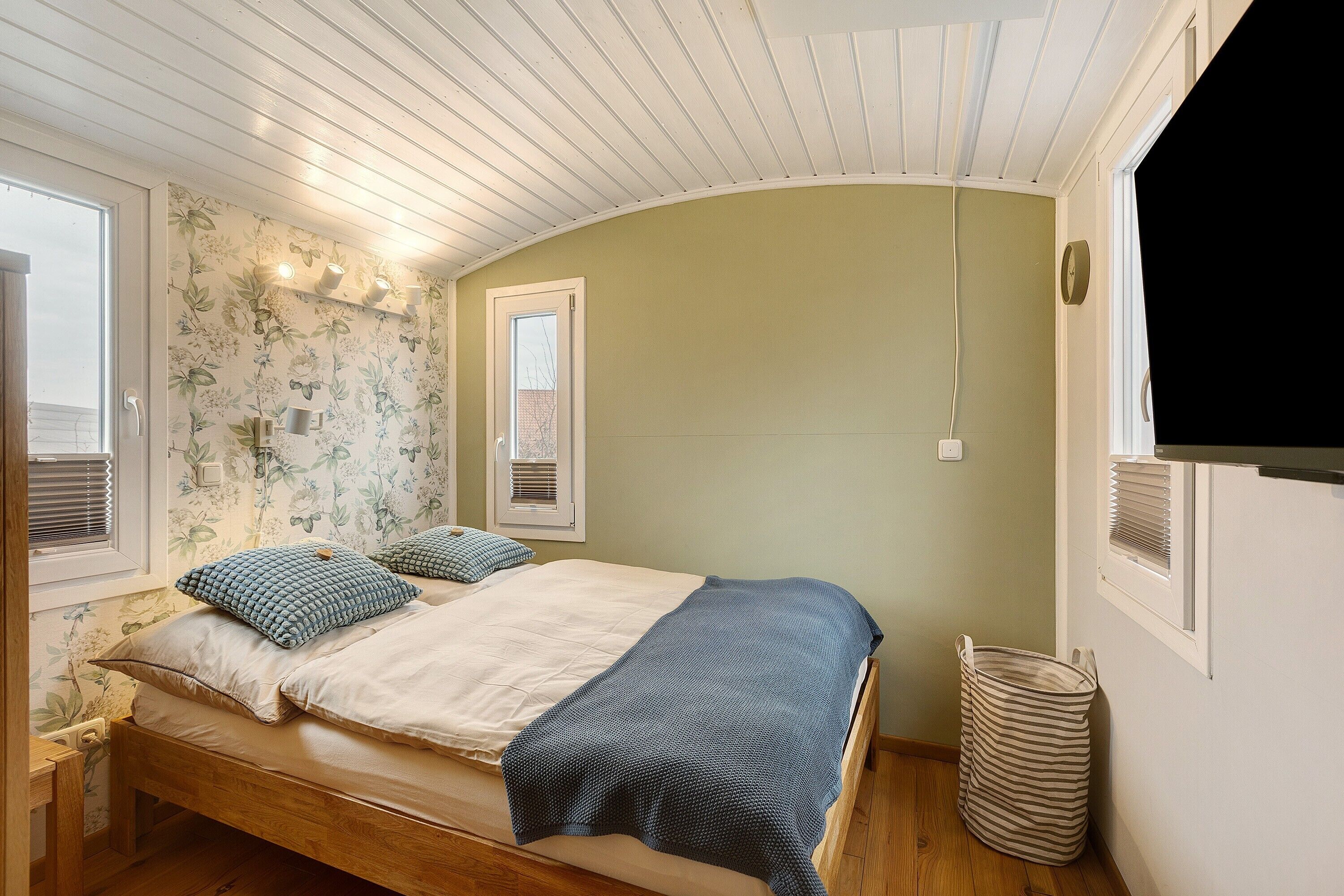 A bedroom with a wooden bed, a window, a wall-mounted shelf, and a wall-mounted light fixture.