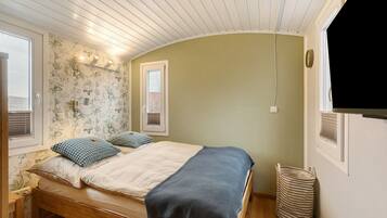 A bedroom with a wooden bed, a window, a wall-mounted shelf, and a wall-mounted light fixture.