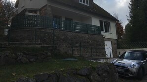 Una casa de dos pisos con cimientos de piedra, un balcón con barandilla verde y un coche estacionado en la parte delantera.