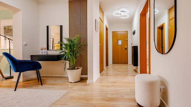 Un pasillo con un mostrador de recepción, una silla azul, una planta en maceta, un reposapiés blanco y una puerta de madera.