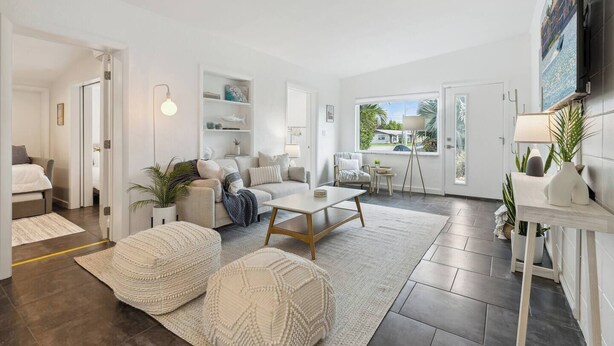 A modern living room with a sofa, coffee table, and ottomans, a dining area with a table and chairs, and a large window offering a view of the outdoors. - Welcome to Space Coast Retreat (Cocoa Beach)