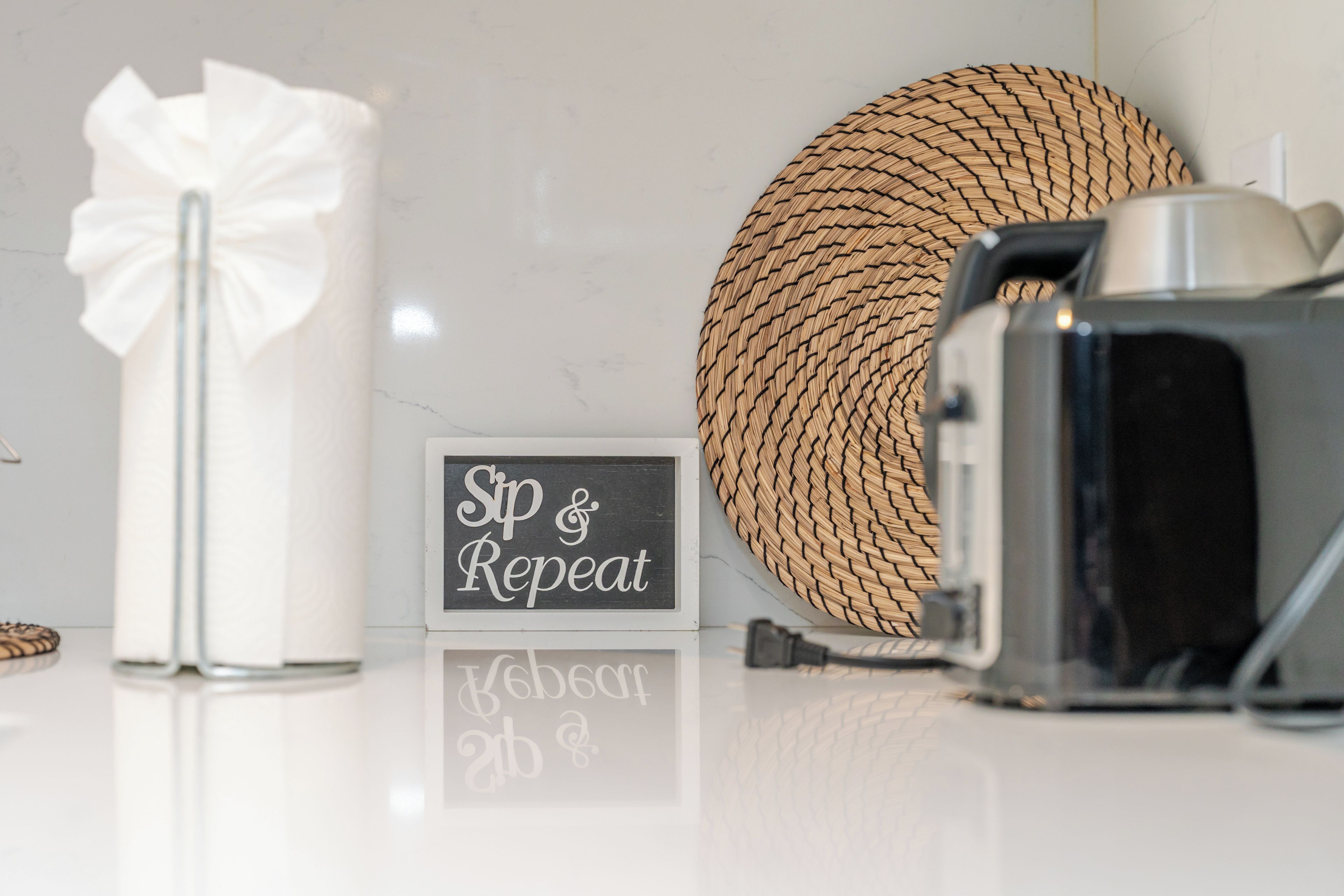 A white table with a black sign reading 'Sip & Repeat,' a white candle, a woven basket, and a vintage camera.