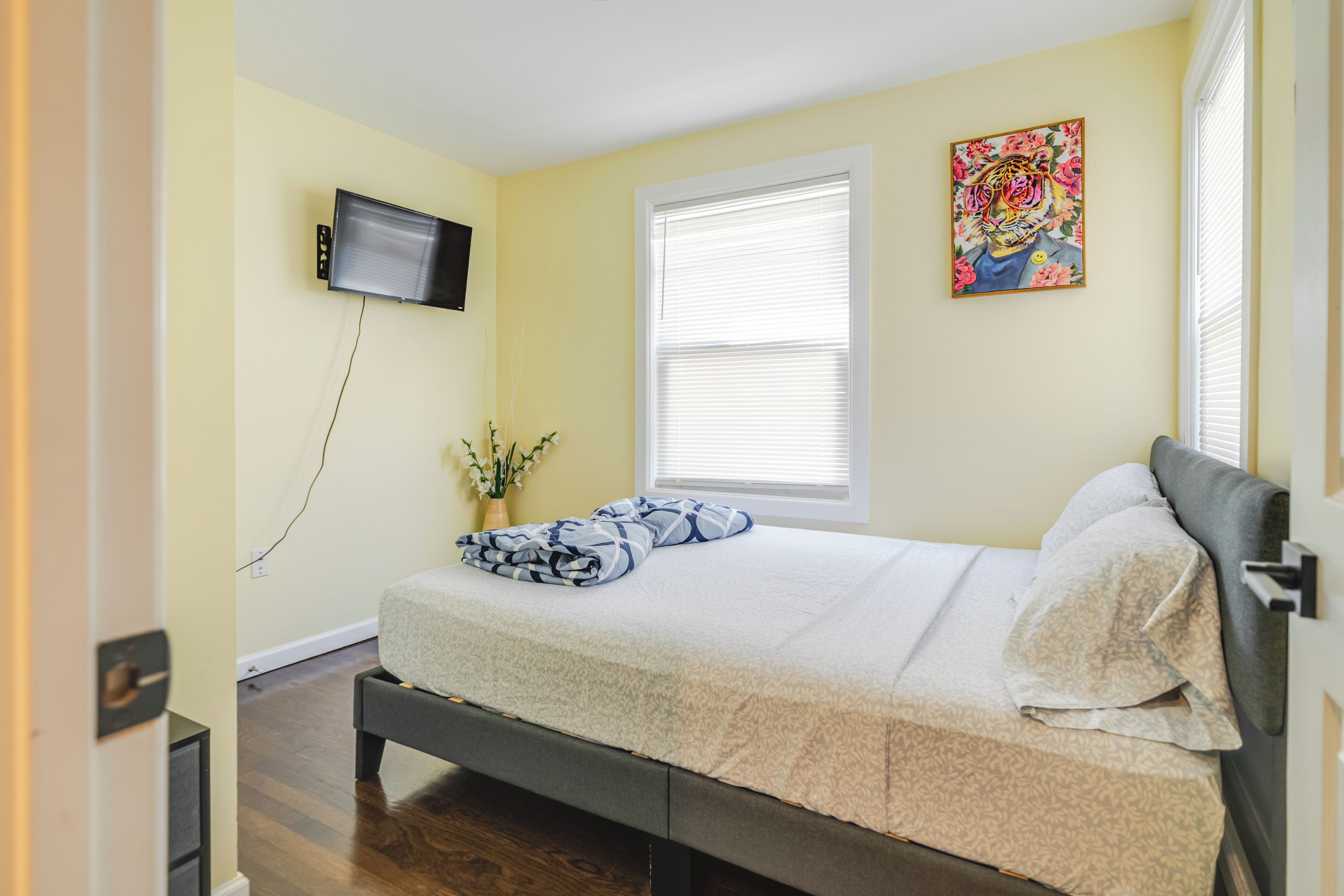 Un dormitorio con cama, televisor montado en la pared, ventana con persianas y un cuadro en la pared.