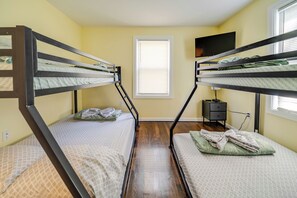A room with bunk beds, a window with blinds, a television on the wall, and a small nightstand.