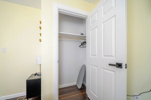 A room with a closed door, a small table with a lamp, a hanging coat rack, and an ironing board.