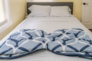 Un lit avec une couette et des oreillers à motifs, une fenêtre avec des stores et une porte en arrière-plan.