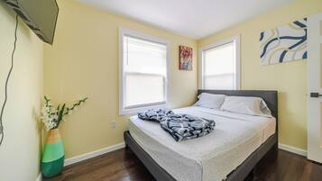 A bedroom with a bed, two windows, a painting, and a vase with flowers.