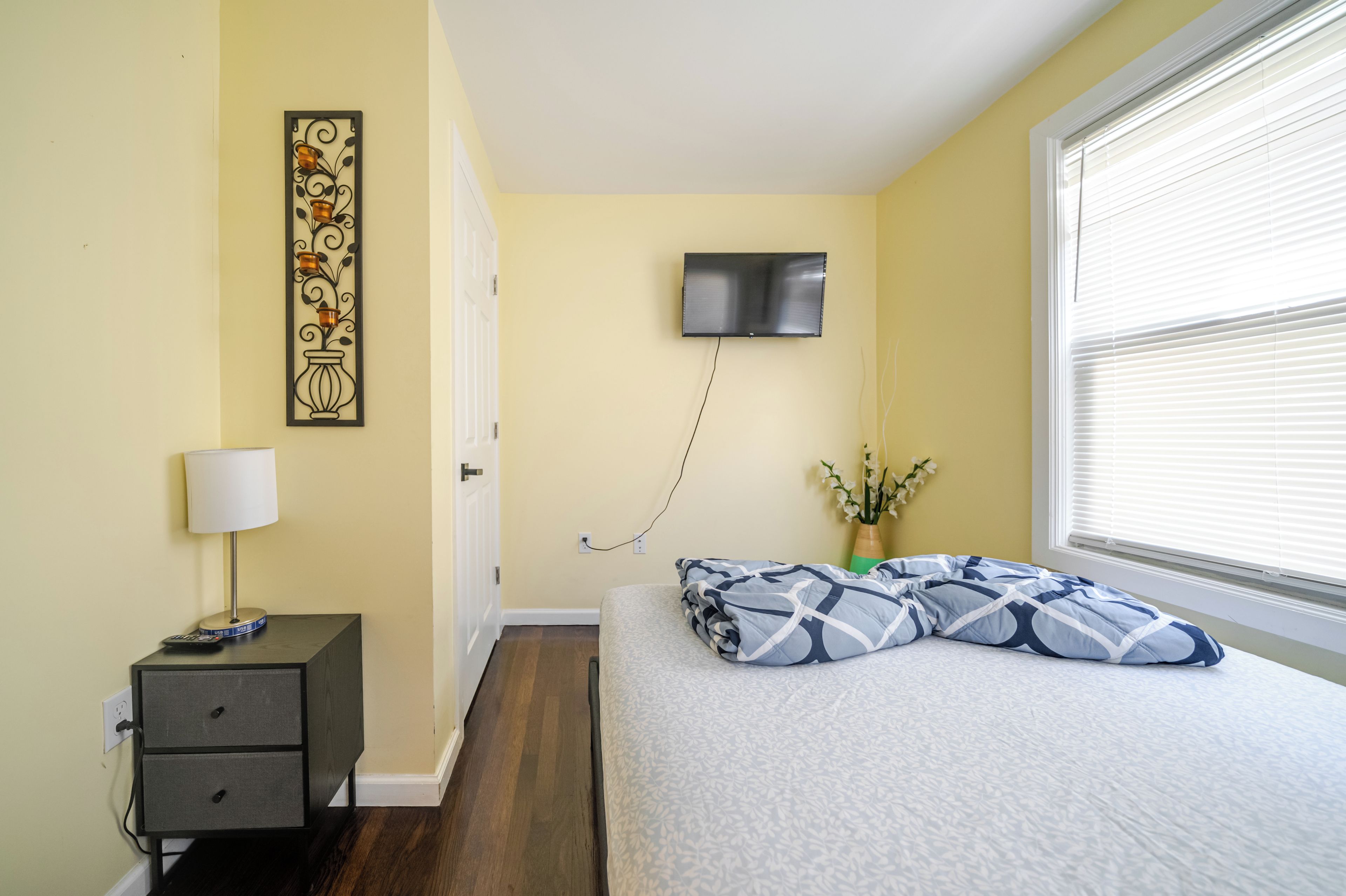 Un dormitorio con cama, una mesita de noche con lámpara, un televisor en la pared y una ventana con persianas.