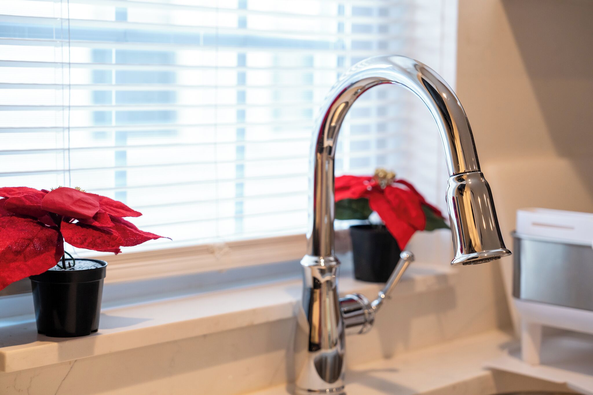 A bathroom with a modern chrome faucet, a potted red plant, and a window with blinds.