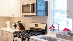 Une cuisine avec des meubles blancs, une cuisinière en acier inoxydable, un four à micro-ondes et une fenêtre avec des stores.