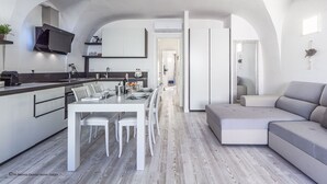 A modern kitchen with white cabinetry, a dining area with a table and chairs, and a grey sofa in a living area. - Gardenia Luxury (Palau)