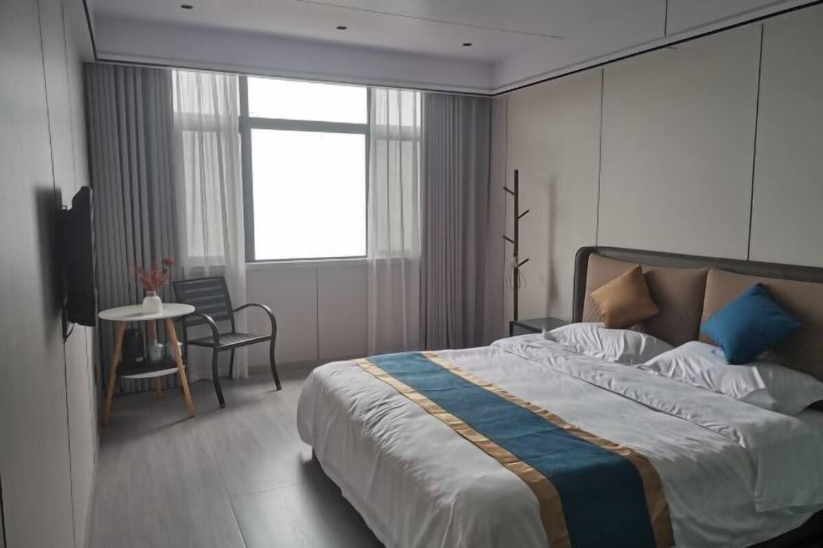 A hotel room with a bed, a chair, a small table, a TV, and a window with curtains.
