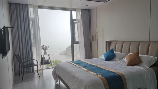 A modern hotel room with a large bed, a small table with a vase, and a view of the sea through the window. - Golden Bay 38 Homestay (Cangnan)