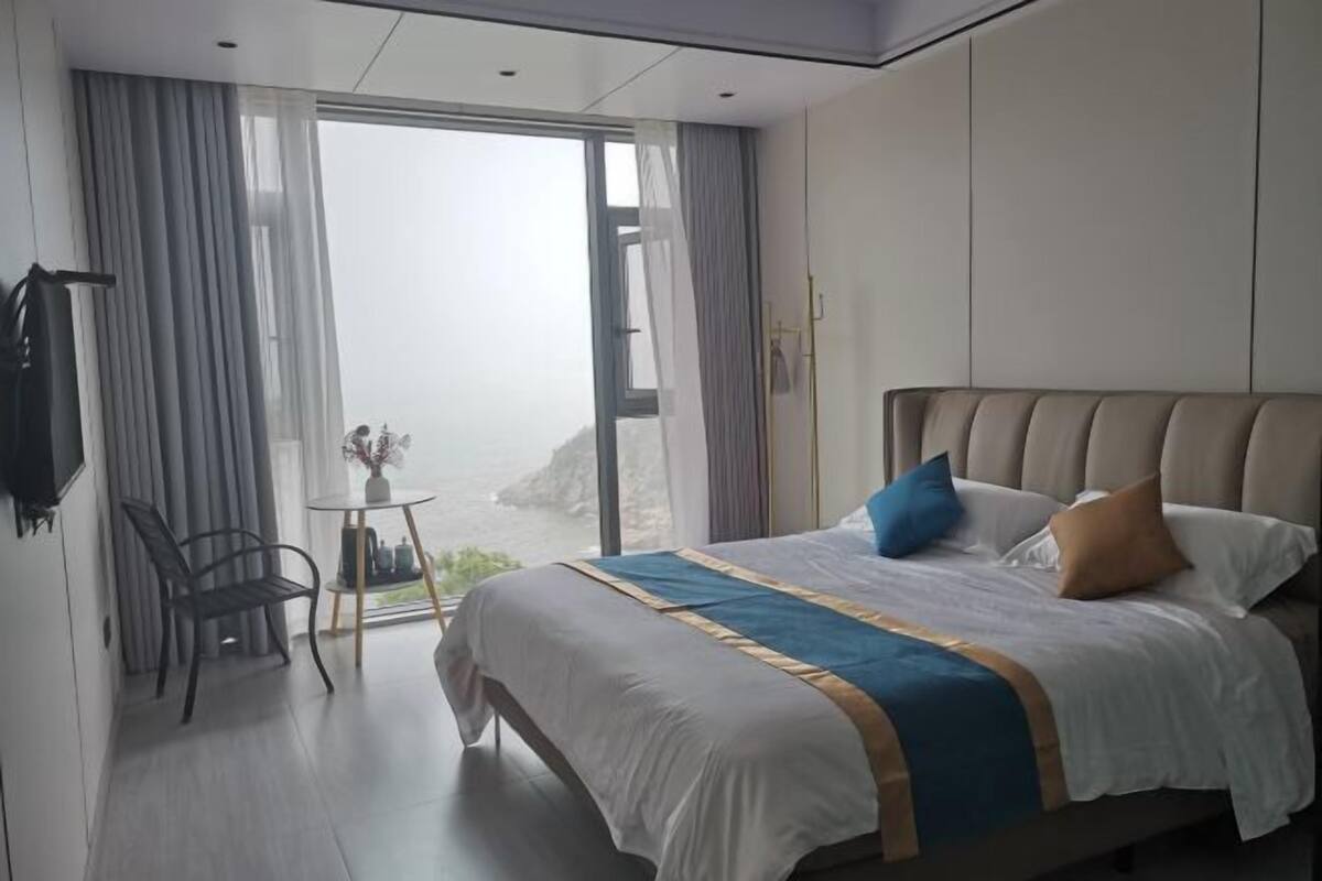 A modern hotel room with a large bed, a small table with a vase, and a view of the sea through the window.