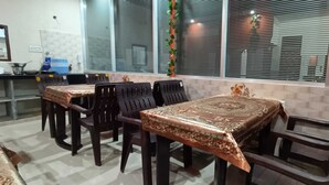 A dining area with tables and chairs, a kitchen area with a stove and sink, and large windows with blinds. - Maheshwari Homestay By GRB (Jamwa Ramgarh)