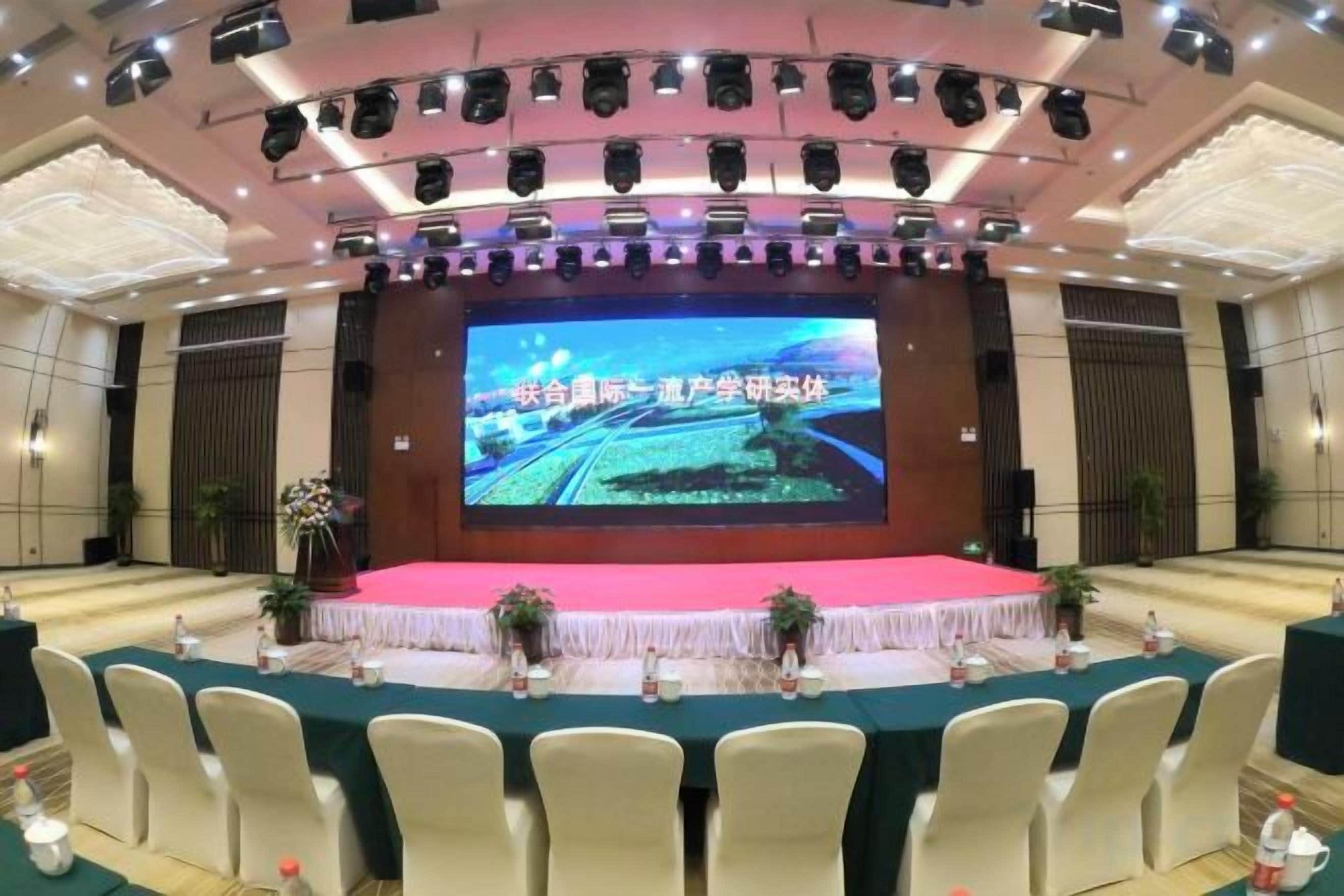 Un salón de conferencias con escenario, una pantalla grande y filas de sillas.