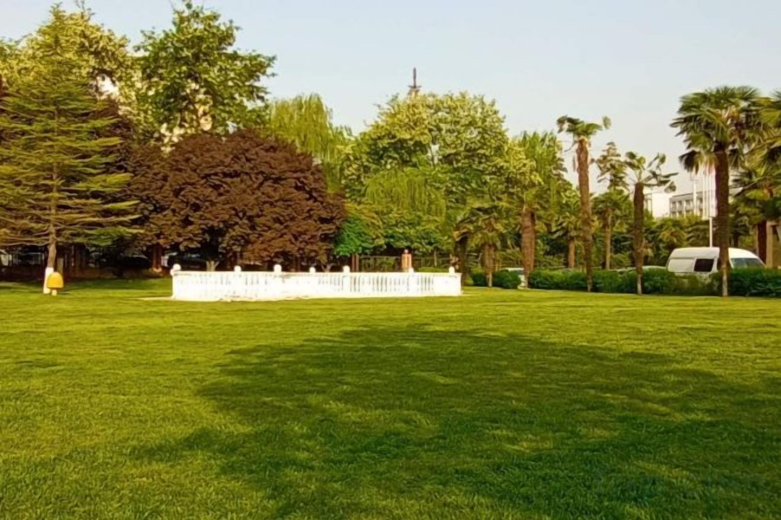 A well-maintained grassy area with a white fence, trees, and a parked vehicle.